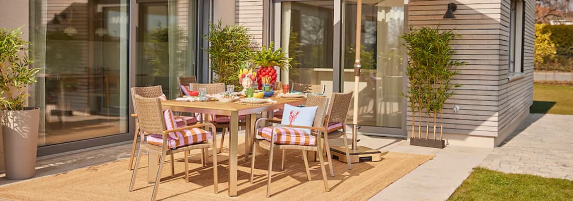 Gedeckter Gartentisch mit sechs Stühlen auf Terrasse vor Haus, bunte Kissen, Geschirr und Pflanzen im Hintergrund