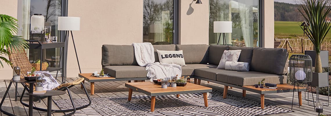 Große Ecklounge auf Terrasse mit Kissen „LEGEND“, niedrigen Tischen, Teppich und Pflanzen vor moderner Hausfassade