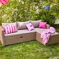 Eckige Gartenlounge mit beigen Polstern, rosa Kissen und Decke auf Rasen vor dichtem grünen Busch