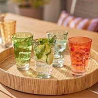 Bunter Mix aus vier Trinkgläsern mit Getränken und Eiswürfeln auf einem runden Tablett auf einem Holztisch draußen