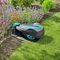 Mähroboter fährt am Rand eines Rasens entlang, daneben blühende Beete und Sträucher im Garten