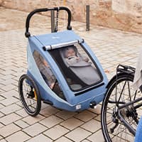 Blauer Fahrradanhänger mit zwei kleinen Kindern im Inneren, angekuppelt an ein Fahrrad auf gepflasterter Straße
