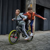 Zwei Kinder fahren gemeinsam auf einem BMX-Rad durch eine Gasse, das Mädchen lenkt, der Junge steht hinten mit ausgebreiteten Armen