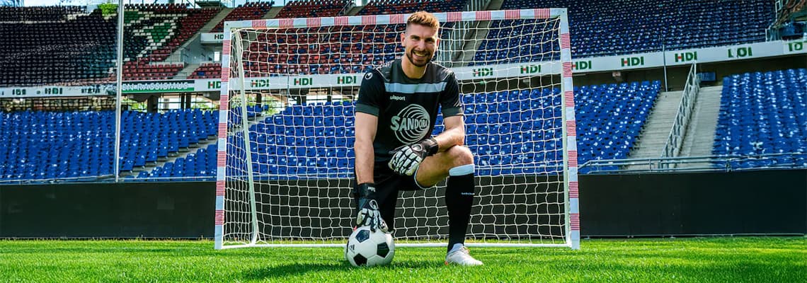 Torwart im schwarzen Trikot kniet vor dem Tor im Stadion und hält einen Fußball auf dem Rasen