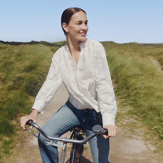 Frau in weißer Bluse und Jeans fährt Fahrrad auf einem sandigen Weg durch eine grüne Dünenlandschaft
