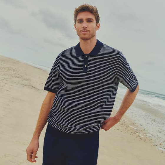 Mann im dunkel gestreiften Poloshirt steht mit einer Hand in der Hüfte am Strand nahe der Wasserlinie