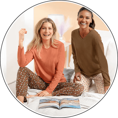 Zwei Frauen im Bett in langärmligen Oberteilen und gemusterten Schlafhosen, vor ihnen liegt eine geöffnete Zeitschrift
