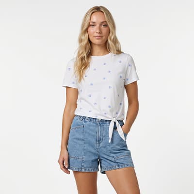 Frau mit langen Haaren trägt weißes T-Shirt mit Knoten vorn und blauen Mustern zu locker sitzenden Jeansshorts