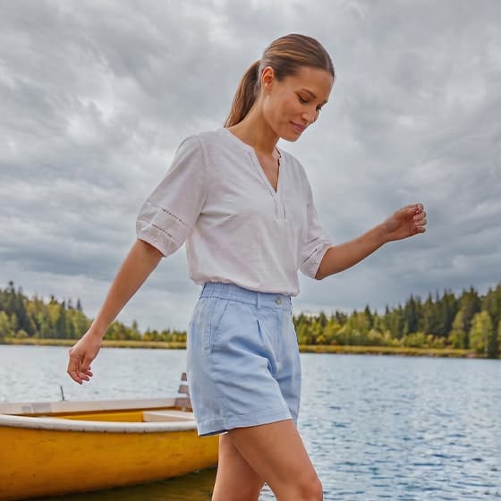 Frau in weißer Bluse und hellblauen Shorts spaziert am Seeufer, im Hintergrund ein gelbes Ruderboot und Wald