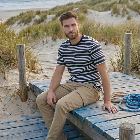 Mann im gestreiften T-Shirt und Chinohose sitzt entspannt auf Holzsteg in den Dünen mit Blick aufs Meer