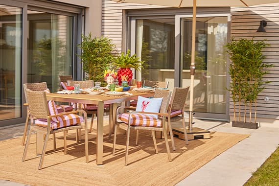 Esstischgruppe auf Terrasse, gedeckter Tisch mit Geschirr und Obst, Stühle mit karierten Kissen auf großem Outdoor-Teppich