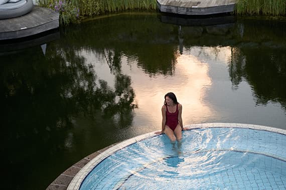 Frau im Badeanzug sitzt am Rand eines runden Pools vor dunklem Naturteich mit Spiegelung des Himmels