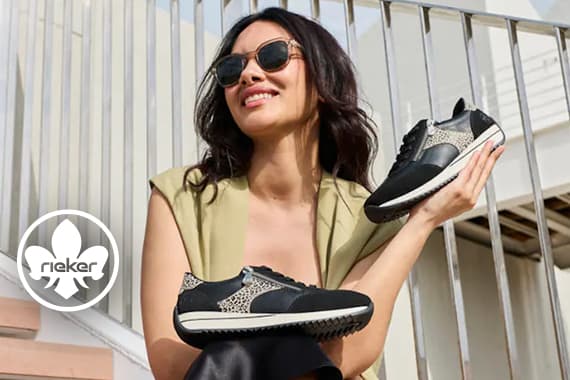 Rieker Logo – Frau mit Sonnenbrille sitzt auf Treppe und hält ein Paar schwarz gemusterte Sneaker in den Händen