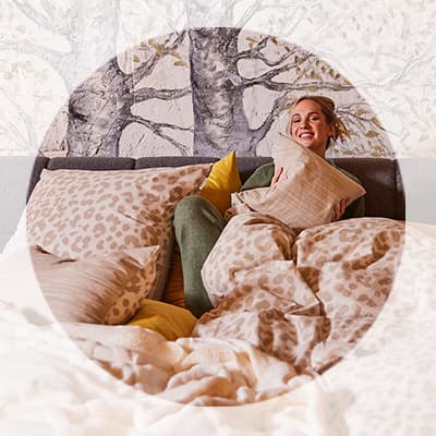 Frau sitzt lachend im Bett, umgeben von gemusterter Bettwäsche und Kissen in Beige- und Gelbtönen