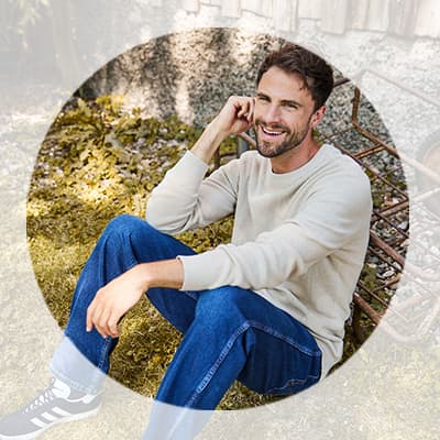 Mann in hellem Pullover und Jeans sitzt entspannt im Gras und lächelt in die Kamera
