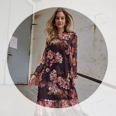 Frau mit langen Haaren trägt knielanges Kleid mit großem Blumenmuster und blickt in einem hellen Flur in die Kamera