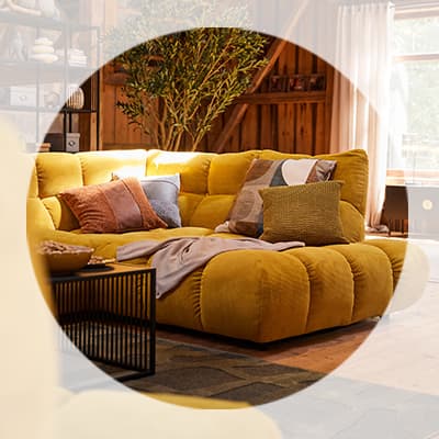 Gelbes Sofa mit vielen Kissen und Decke, daneben schwarzer Beistelltisch in einem hellen Wohnzimmer mit großen Fenstern