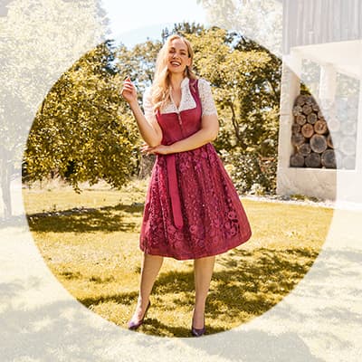 Frau im pinken Dirndl mit Spitzenbluse steht lächelnd im sonnigen Garten vor Bäumen und einem Holzstapel