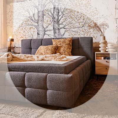 Gepolstertes Doppelbett mit Kissen und Decke, daneben Nachttisch mit Lampe und Deko vor Wand mit Baum-Motiv