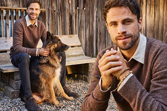 Mann im braunen Pullover sitzt mit Schäferhund auf Bank vor Holzwand, Nahaufnahme seines Gesichts rechts im Bild