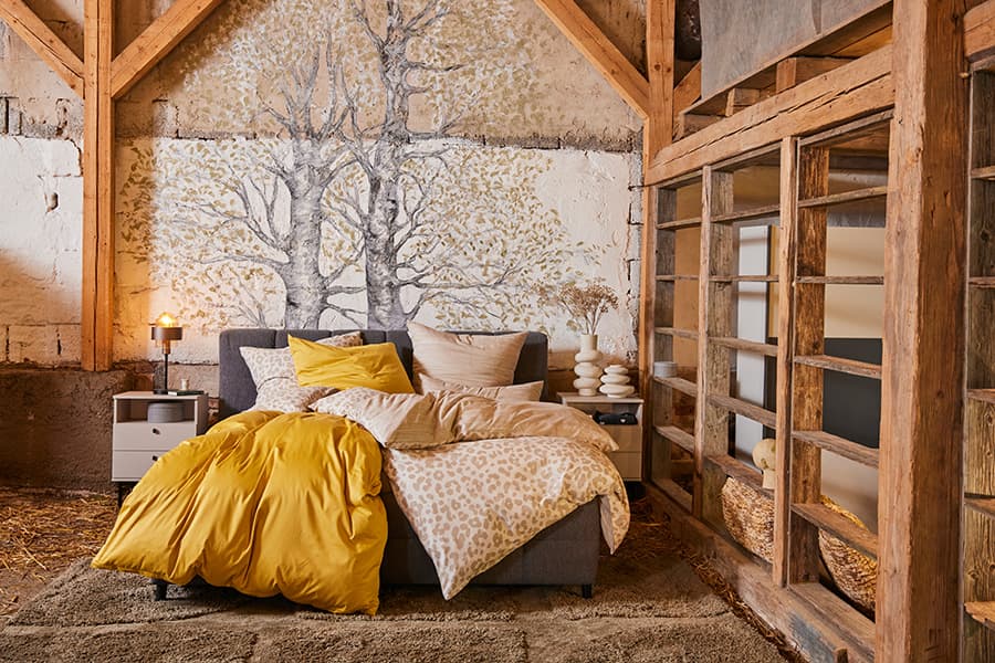 Doppelbett mit gelb-gemusterter Bettwäsche vor Wandbild mit Baum, eingerahmt von Holzbalken und Nachttischen mit Lampen
