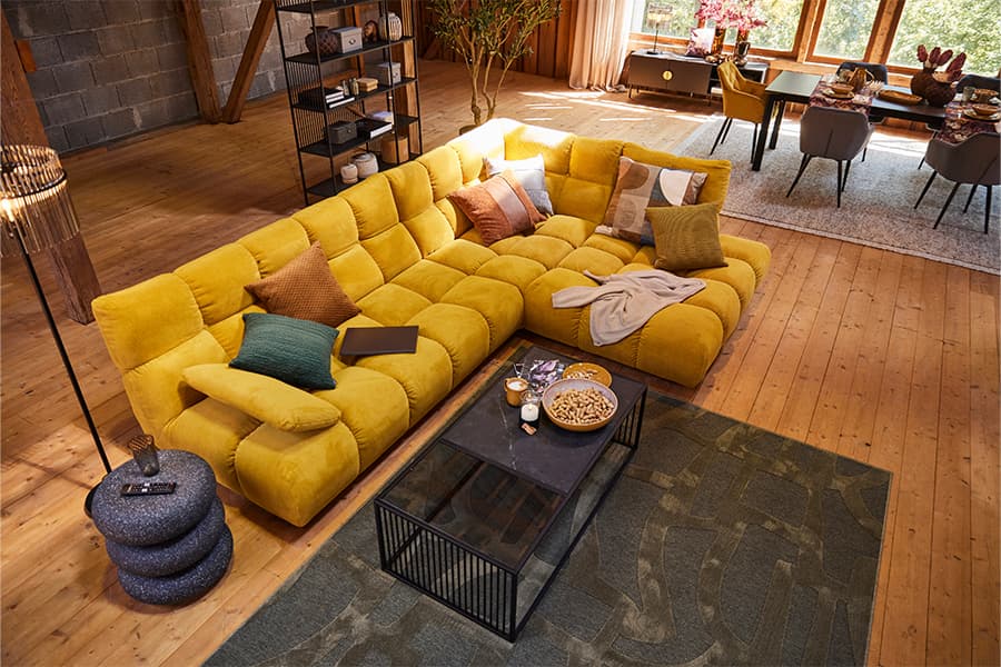 Großes gelbes Ecksofa mit vielen Kissen in modernem Wohnzimmer, davor Couchtisch mit Snacks und Kerzen