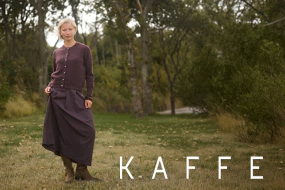 KAFFE – Frau im langen dunklen Rock und passender Strickjacke steht auf einer Wiese vor Bäumen