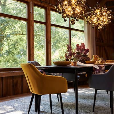 Essbereich mit gelbem und grauem Stuhl an dunklem Tisch, große Fenster mit Blick ins Grüne und leuchtende Deckenlampen