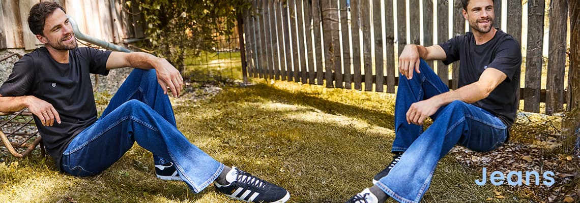 Jeans – Mann in blauem Jeans und schwarzem T-Shirt sitzt entspannt im Garten vor Holzzaun