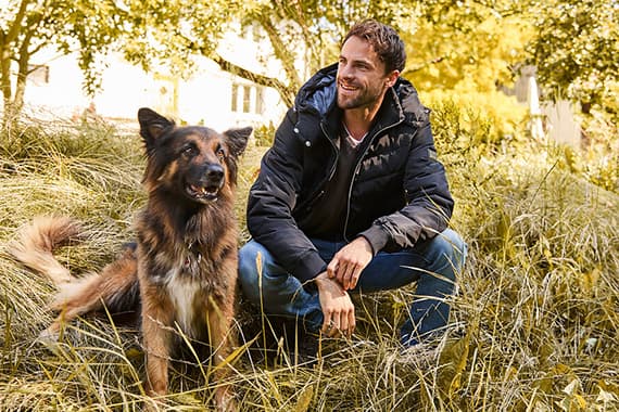 Mann in dunkler Jacke hockt im hohen Gras und schaut seinen neben ihm sitzenden Hund im Park an