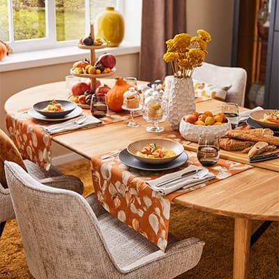 Herbstlich dekorierter Esstisch mit orangefarbenen Tischläufern, gedeckten Tellern, Obstschalen und Blumenvasen.