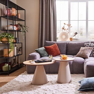 Modernes Wohnzimmer mit grauem Sofa, bunten Kissen, zwei Beistelltischen und Regal mit Pflanzen und Dekoartikeln