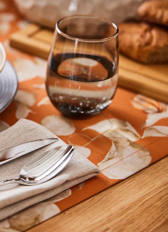 Wasserglas, Besteck und Brote auf einem orangefarbenen Tischläufer mit weißen Blättern, Tischgedeck mit Serviette