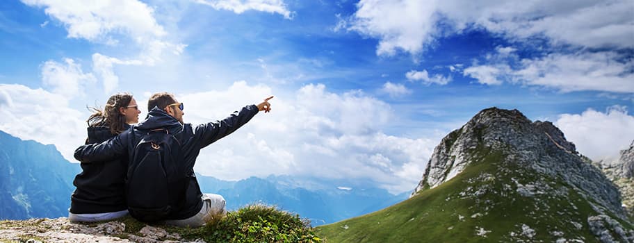 Zwei Personen mit Rucksäcken sitzen auf einem Berg und schauen in die Ferne, eine Person zeigt auf einen Berggipfel unter blauem Himmel
