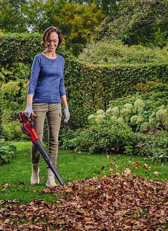 Frau in blauem Langarmshirt und beigen Hosen nutzt Laubbläser im Garten, steht neben einem Haufen Herbstlaub