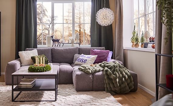 Wohnzimmer mit grauem Sofa, Kissen mit Aufschrift "Alles ganz leise", Adventskranz, Fensterdekoration und Pflanzen auf Fensterbank