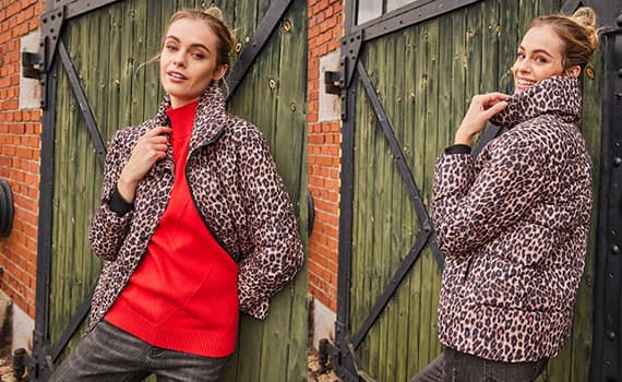 Frau trägt eine Leoparden-Jacke über einem roten Pullover, steht vor grüner Holztür und Backsteinwand, Gesicht unscharf.