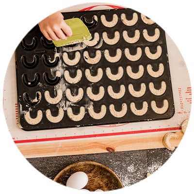 Blech mit ungebackenen Plätzchen in Hufeisenform, Hand verteilt Teig, darunter Schüssel mit Eiern und Teigrolle