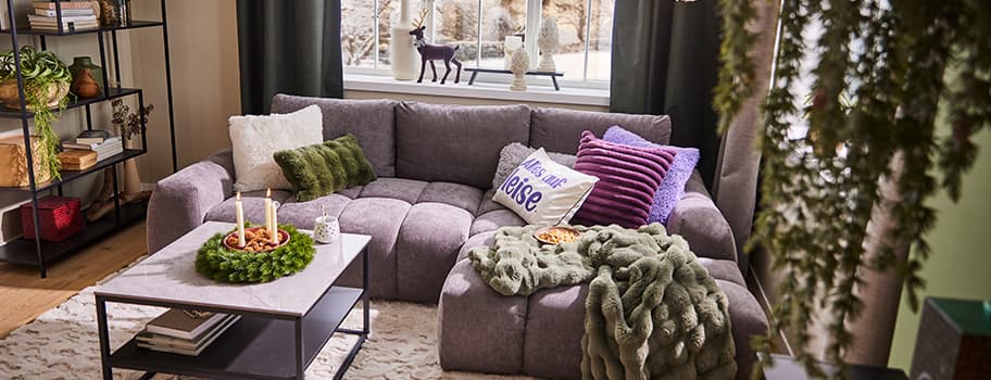 Wohnzimmer mit grauem Sofa, grüner Decke, Kerzen auf Tisch, Kissen mit "Reise" und Fenster mit Dekofiguren im Hintergrund