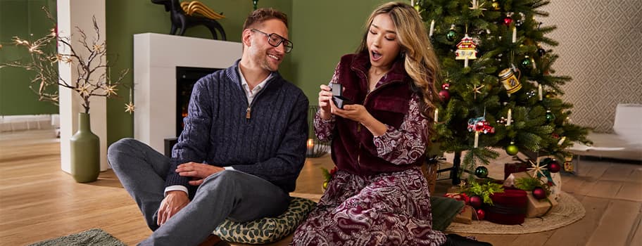 Paar sitzt vor Weihnachtsbaum und Kamin, Frau zeigt Geschenkbox mit Schmuckstück, Mann schaut zu, festliche Dekoration im Hintergrund