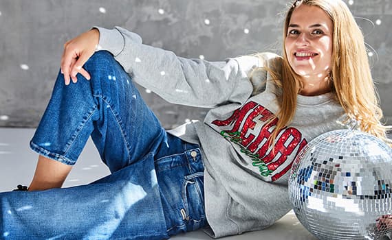 Frau in grauem Sweatshirt mit rotem Schriftzug und blauer Jeans liegt neben einer silbernen Discokugel