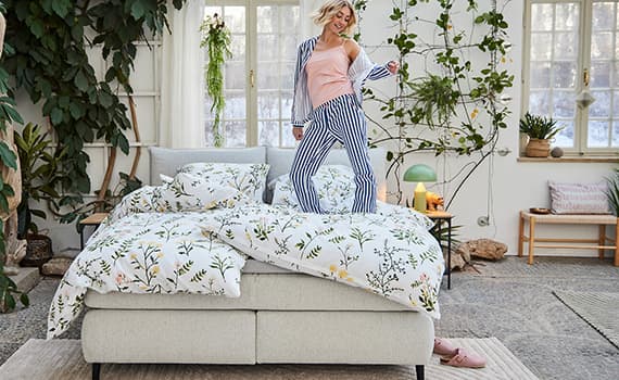 Person in gestreifter Schlafanzughose steht auf einem Bett mit floraler Bettwäsche in einem hellen Raum mit Pflanzen.