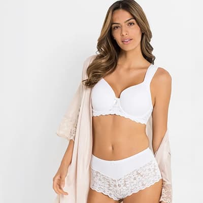 Frau in weißem Spitzen-BH und passendem Höschen, trägt einen beigen Morgenmantel.