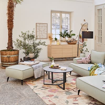 Modernes Wohnzimmer mit grüner Couch, Teppich, Pflanzen und Holzschränken, Tageslicht durch Fenster.