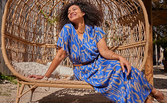 Frau in blau gemustertem Kleid sitzt entspannt in einem Korbsessel im Freien